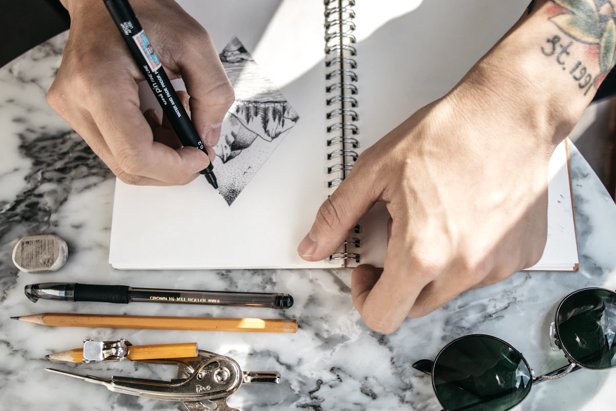 Mãos de um designer a desenhar esboços de um logótipo num caderno, com canetas e lápis sobre a mesa