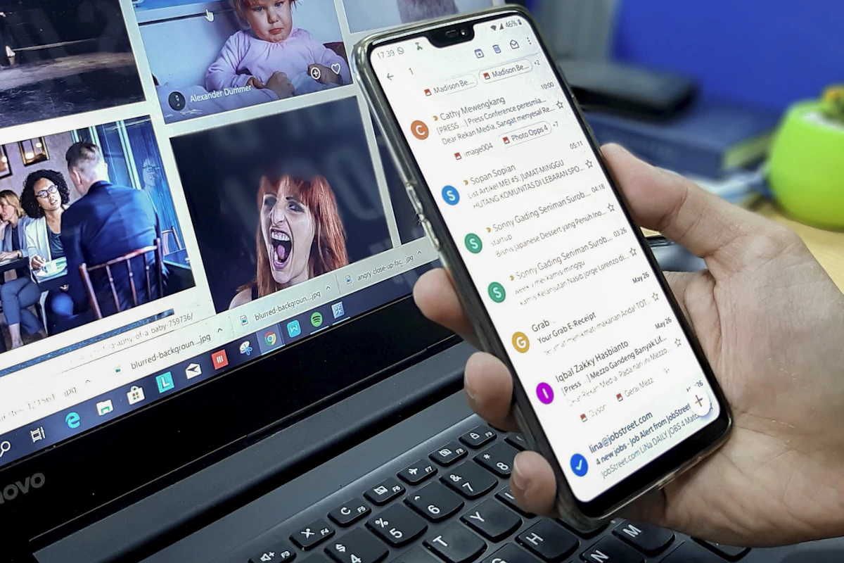 Mão a segurar um telemóvel com uma lista de emails aberta, em frente a um portátil