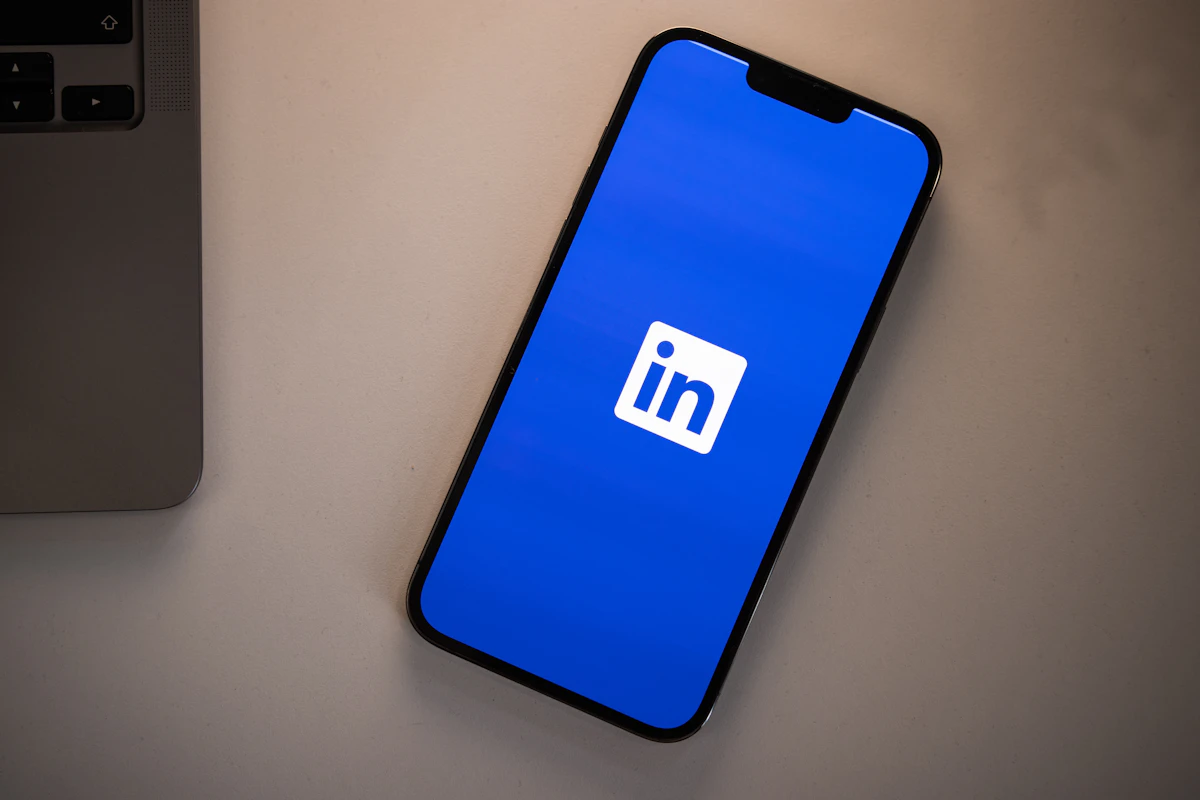 Telemóvel com o logótipo do LinkedIn no ecrã azul, pousado numa secretária ao lado de um portátil