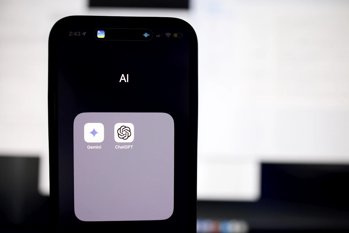 Mão a segurar um telemóvel com uma pasta de apps de IA aberta, onde aparecem o Gemini e o ChatGPT