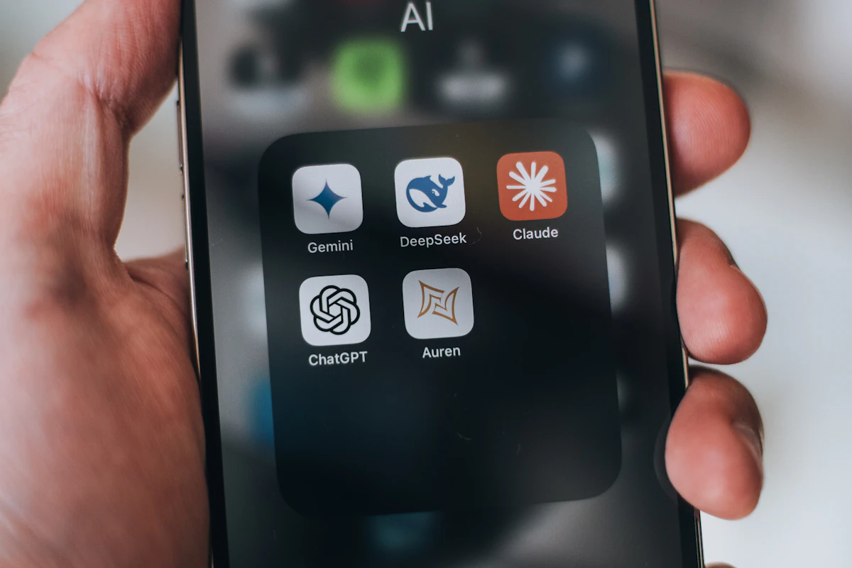 Mão a segurar um telemóvel com uma pasta de apps de IA, entre elas Gemini, DeepSeek, Claude e ChatGPT