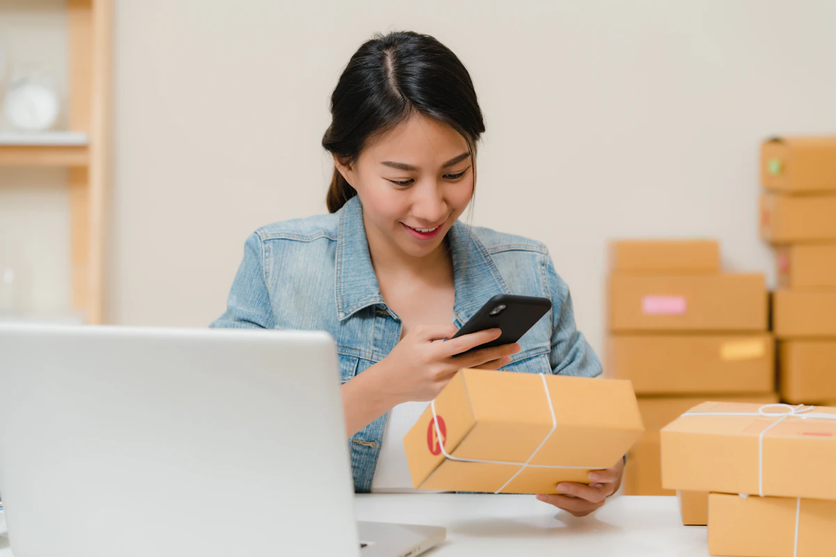 Mulher a verificar uma encomenda com o telemóvel na mão, rodeada de caixas e de um portátil